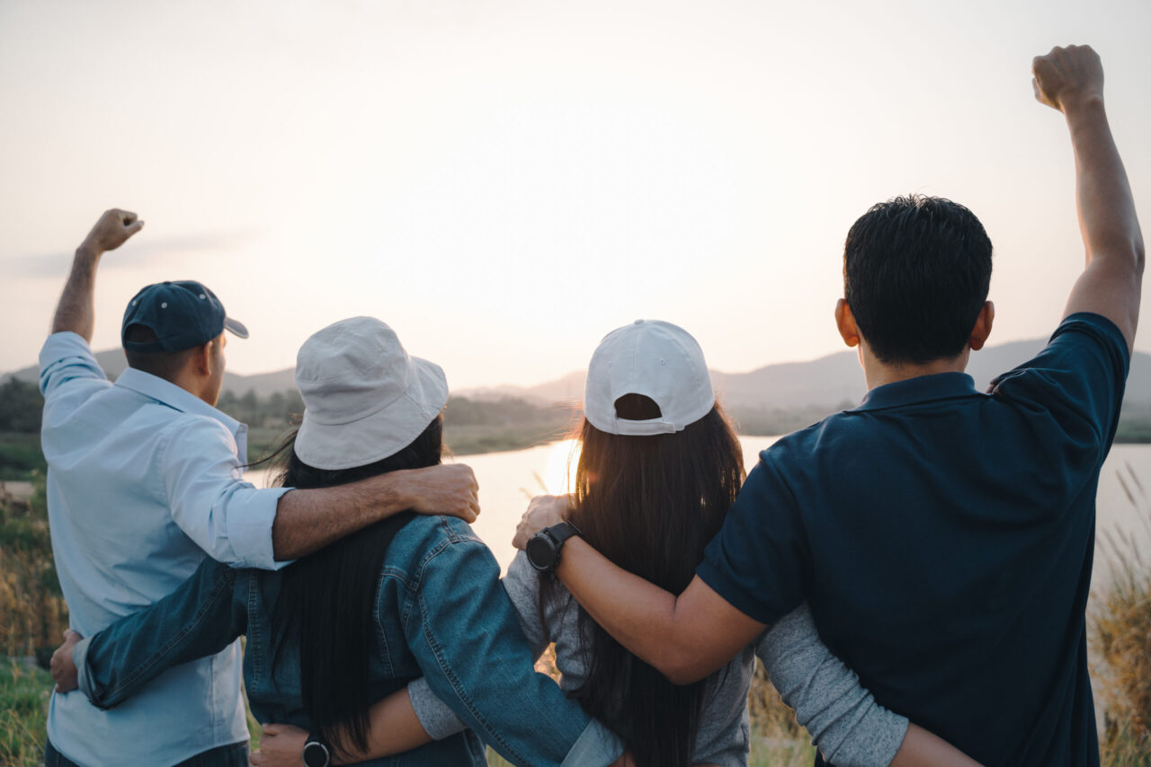 group of people celebrating with raised arms looking at sunrise on the mountain background.